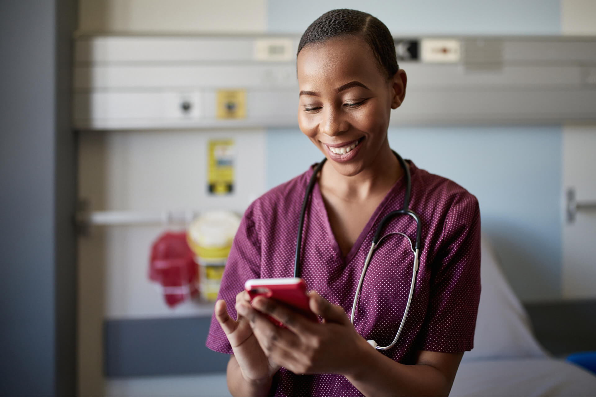 About Us - Discover Healthcare Careers Young Black nurse in purple scrubs wearing stethoscope scrolling on smartphone
