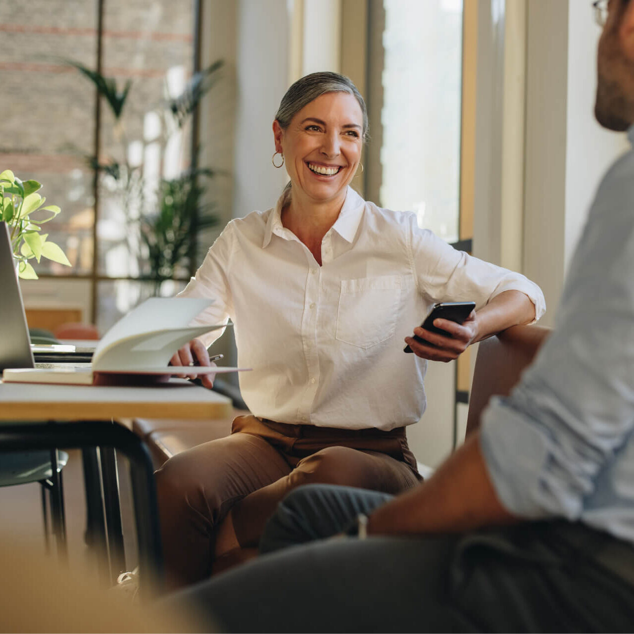 Corporate Careers - Encouraging Culture Middle-aged businesswoman holding smartphone and talking to coworker in office setting